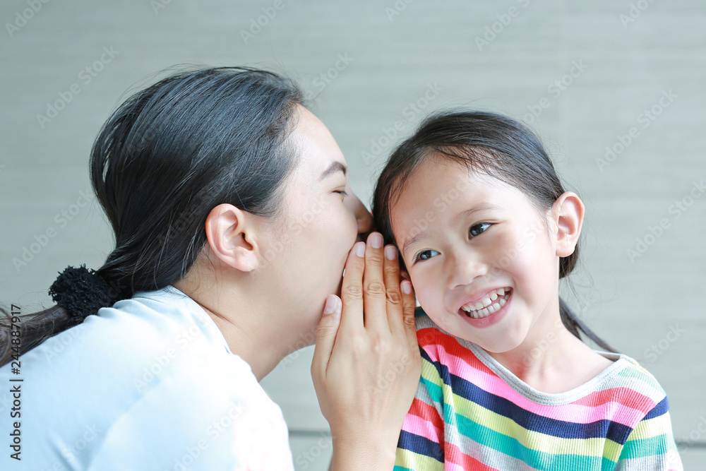 Portrait of happy mom whispering a something secret to her little daughter ear on sofa at home. Mother and kid communication concept. Portrait of happy mom whispering a something secret to her little daughter ear on sofa at home. Mother and kid communication concept.