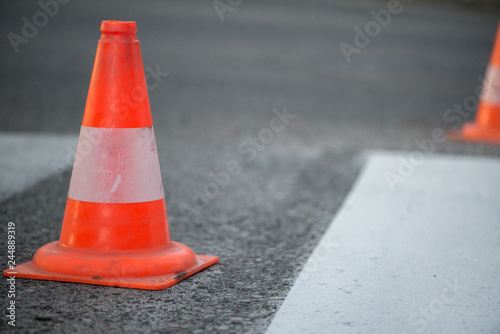 Macro shot of road traffic cones with orange and white stripes standing on street on gray asphalt during road construction works. Just painted white street lines on pedestrian crossing 