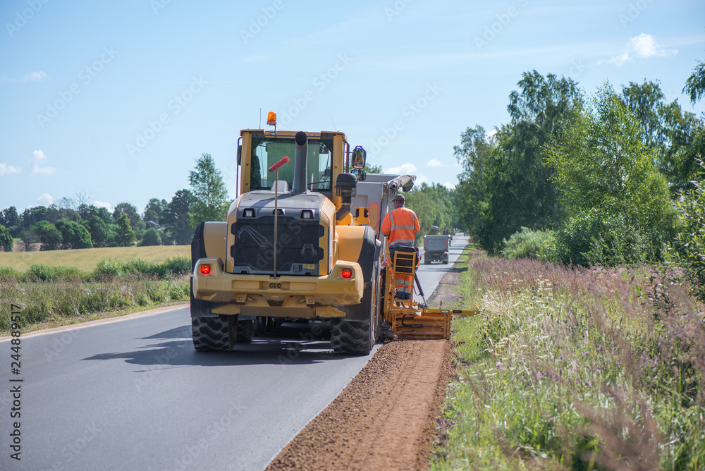 Road construction workers repairing highway road on sunny summer day. Loaders and trucks on newly made asphalt. Heavy machinery working on street. Road curbs being constructed with gravel 