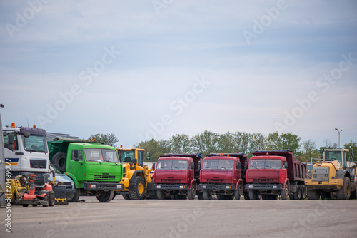 Multiple cars, trucks, loaders, concrete mixers and construction machinery in large parking lot in industrial territory, next to concrete and asphalt factory 