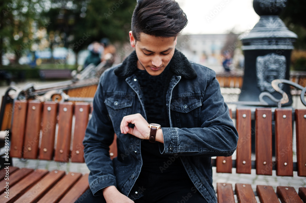 Handsome and fashionable indian man in black jeans jacket posed outdoor,  sitting on bench and looking at his watches. Photos | Adobe Stock