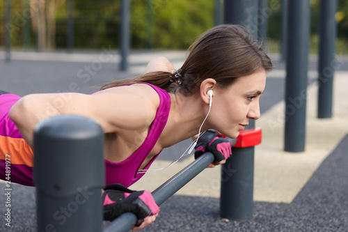 Young caucasian woman in a bright sportswear pushes up in outdoor sportground. White earphones, protective gloves. Summer, spring, early autumn. Real woman in morning training, smiling. Copy space.
