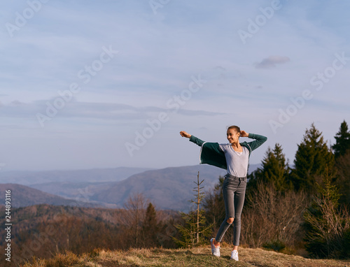 happy woman hiker standing raising hands on edge of mountain ridge against background of mountains and blue sky on sunset