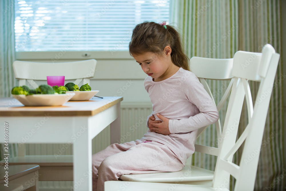 Child sitting at the table in the kitchen with stomach pain. Hands on belly. Little girl suffering Child sitting at the table in the kitchen with stomach pain. Hands on belly. Little girl suffering