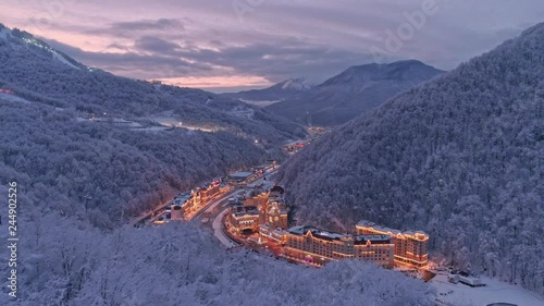 Aerial drone establishing shot of white winter valley Rosa Khutor resort in Sochi. Snow covered trees. Evening illumination. After sunset. Flying low above snow forest