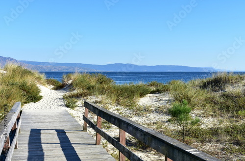 Beach with vegetation in sand dunes and wooden boarwalk with fence. Blue sea  clear sky  sunny day. Galicia  Spain.