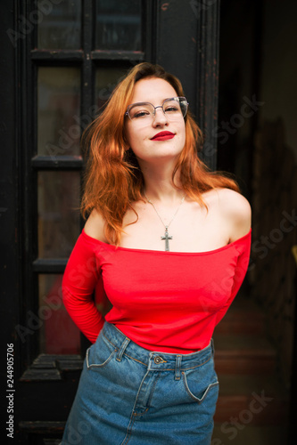 Attractive redhaired woman in eyeglasses, wear on red blouse and jeans skirt posing at street against old wooden door.
