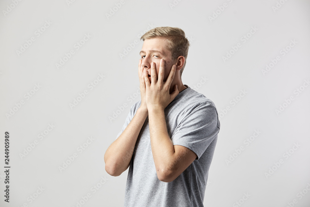 Sad guy dressed in a white t-shirt holds his hands on his face on the white background in the studio