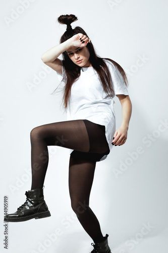 Stylish young dark-haired girl with a funny hairstyle dressed in white t-shirt, black tights and shoes is posing on the white background in the studio