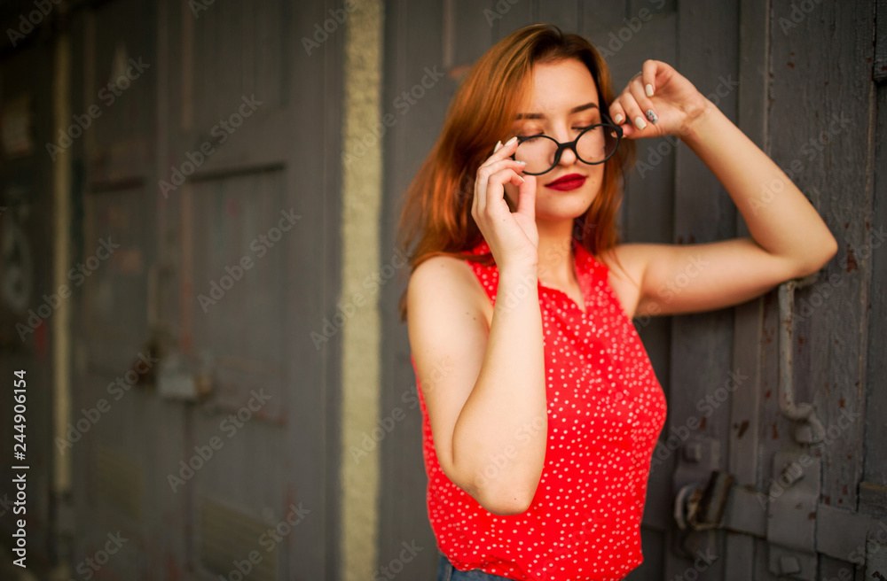 Fototapeta premium Attractive redhaired woman in eyeglasses, wear on red blouse and jeans skirt posing.