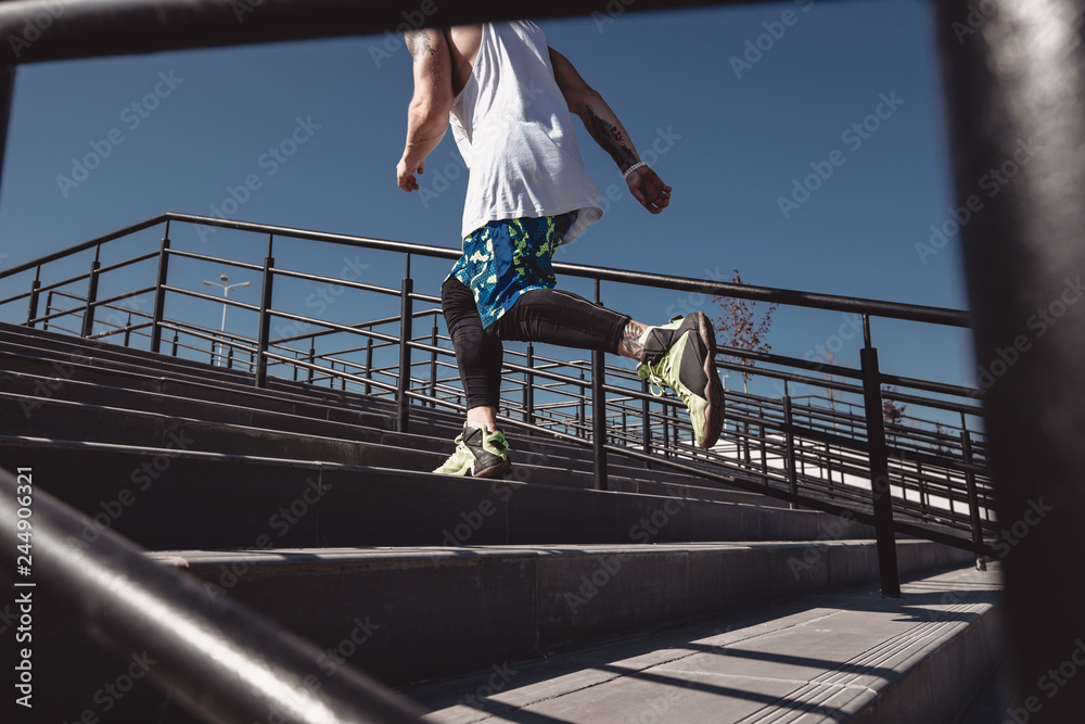 Athletic man with headband on his head dressed in the white t-shirt, black leggings and blue shorts is running up the stairs  with black railings outside on a sunny day