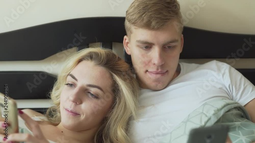 A close up portrait video. A beautiful happy married couple lies in the bed. Each minds his/her own business and checks own phone. The wife glances into her husband's phone with the smile on her face. photo