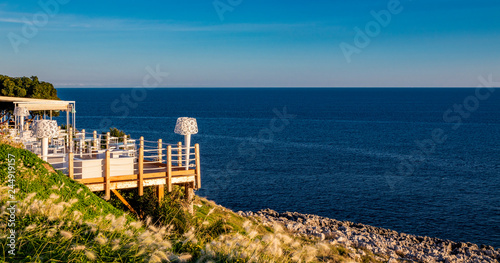 Italian restaurant with panoramic terrace, with spectacular view of the blue sea. A wonderful place in Puglia, Italy. Wooden terrace with curtains, armchairs, chairs, tables and lamps in white.