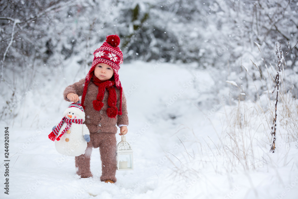 Portrait of a cute toddler baby dressed in a brown hand knitted jacket, pants, red hat and scarf, holding teddy and lantern, walks through the snowy park enjoying first snow blowing
