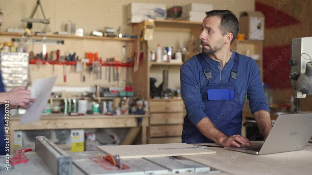 Lockdown of middle-aged joiner using laptop, while his young colleague coming and starting showing some blueprints Lockdown of middle-aged joiner using laptop, while his young colleague coming and starting showing some blueprints