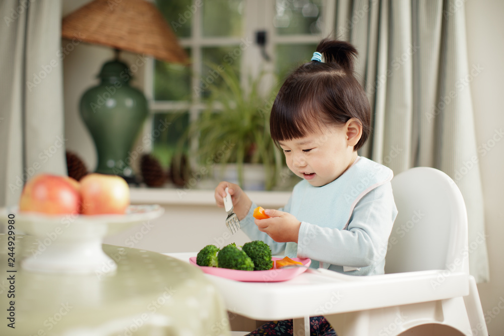 toddler girl eating healthy vegetable sitting on high chair beside a dinner table at home toddler girl eating healthy vegetable sitting on high chair beside a dinner table at home