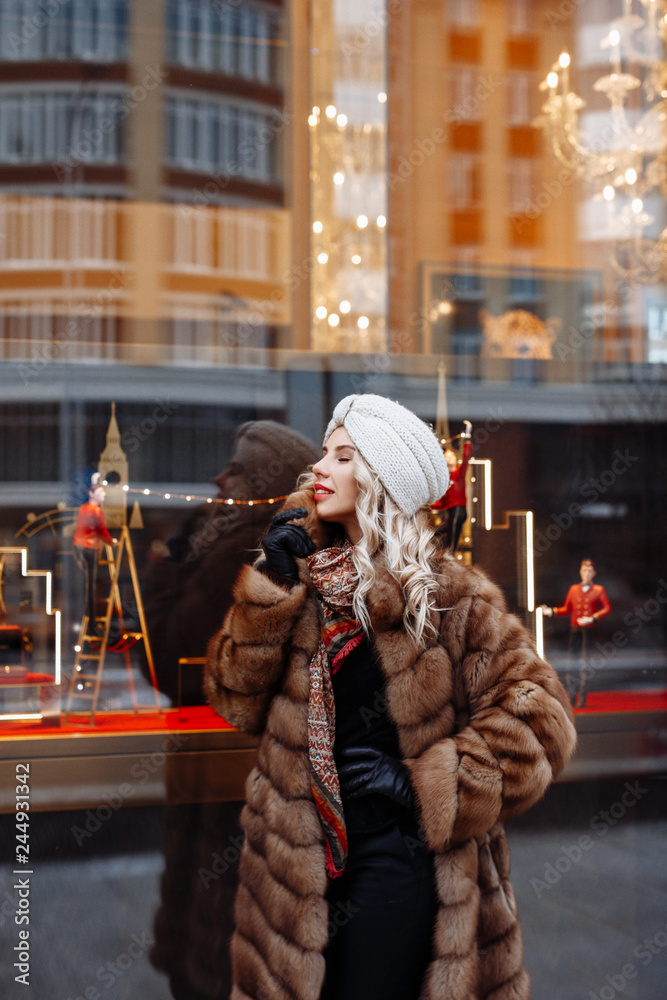 Outdoor close up portrait of beautiful  smiling woman blonde with curly hair, dressed in a white knitted hat and fur coat posing in street of european city. Winter fashion, Christmas holidays concept.