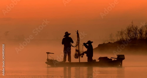 Two silhouetted traditional asian fishermen using nets to fish at an exotic river location from a flat-bottomed boat at sunrise or sunset