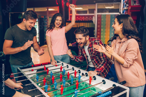 Cheerful happy young team stand at soccer table in playing room and cheering. Bearded guy play with another human. Frieds surround him.
