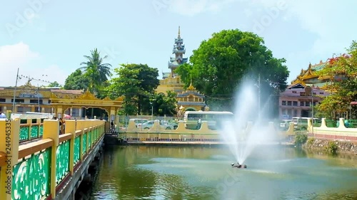 YANGON, MYANMAR - FEBRUARY 17, 2018: Fountain in pond of Mahavijaya (Maha Wizaya) Pagoda with a bridge and pyatthat roof of Kyauk Sein Image House behind the trees, on February 17 in Yangon. photo