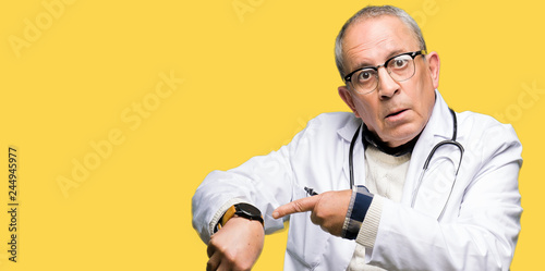 Handsome senior doctor man wearing medical coat In hurry pointing to watch time, impatience, upset and angry for deadline delay