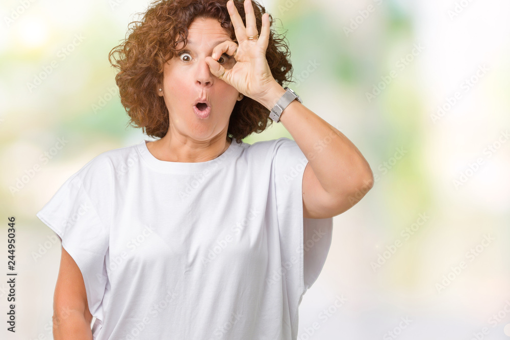 Beautiful middle ager senior woman wearing white t-shirt over isolated background doing ok gesture shocked with surprised face, eye looking through fingers. Unbelieving expression.
