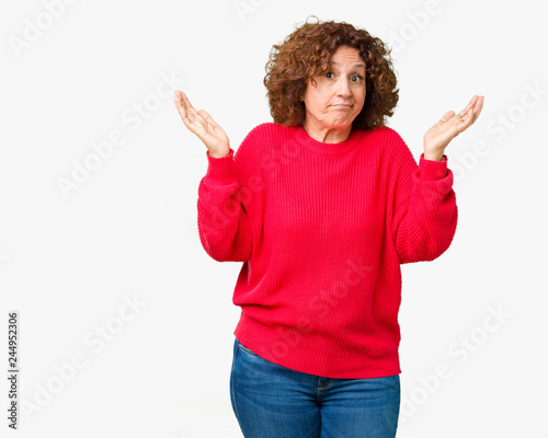 Beautiful middle ager senior woman red winter sweater over isolated background clueless and confused expression with arms and hands raised. Doubt concept.