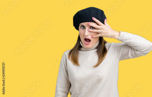 Middle age mature woman wearing winter sweater and beret over isolated background peeking in shock covering face and eyes with hand, looking through fingers with embarrassed expression. © Krakenimages.com