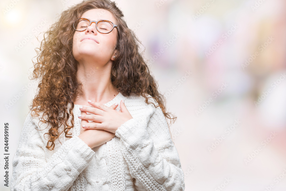 Beautiful brunette curly hair young girl wearing winter sweater over isolated background smiling with hands on chest with closed eyes and grateful gesture on face. Health concept. Beautiful brunette curly hair young girl wearing winter sweater over isolated background smiling with hands on chest with closed eyes and grateful gesture on face. Health concept.