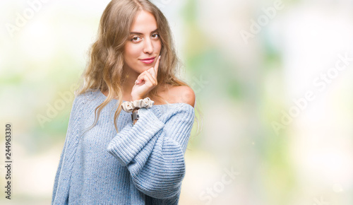 Beautiful young blonde woman wearing winter sweater over isolated background looking confident at the camera with smile with crossed arms and hand raised on chin. Thinking positive.