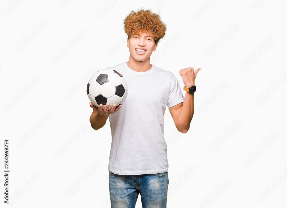 Young handsome man holding soccer football ball pointing and showing with thumb up to the side with happy face smiling