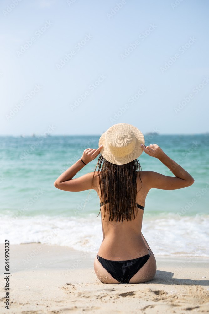 Youg woman in black bikini resting on the beach in straw hat