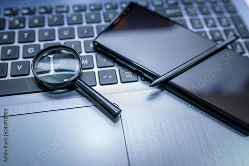 Magnifying glass, modern mobile phone with stylus on laptop computer keyboard in blue tone.