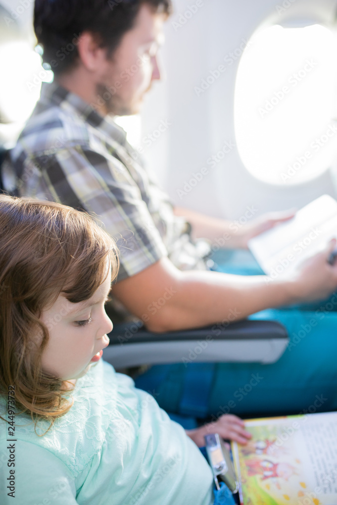 Adorable little girl traveling by an airplane with her father, sitting by aircraft window and reading books Adorable little girl traveling by an airplane with her father, sitting by aircraft window and reading books