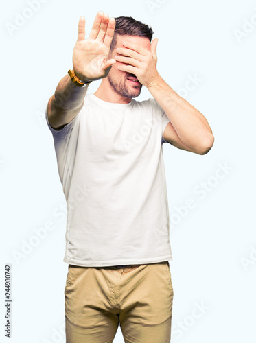 Handsome man wearing casual white t-shirt covering eyes with hands and doing stop gesture with sad and fear expression. Embarrassed and negative concept.