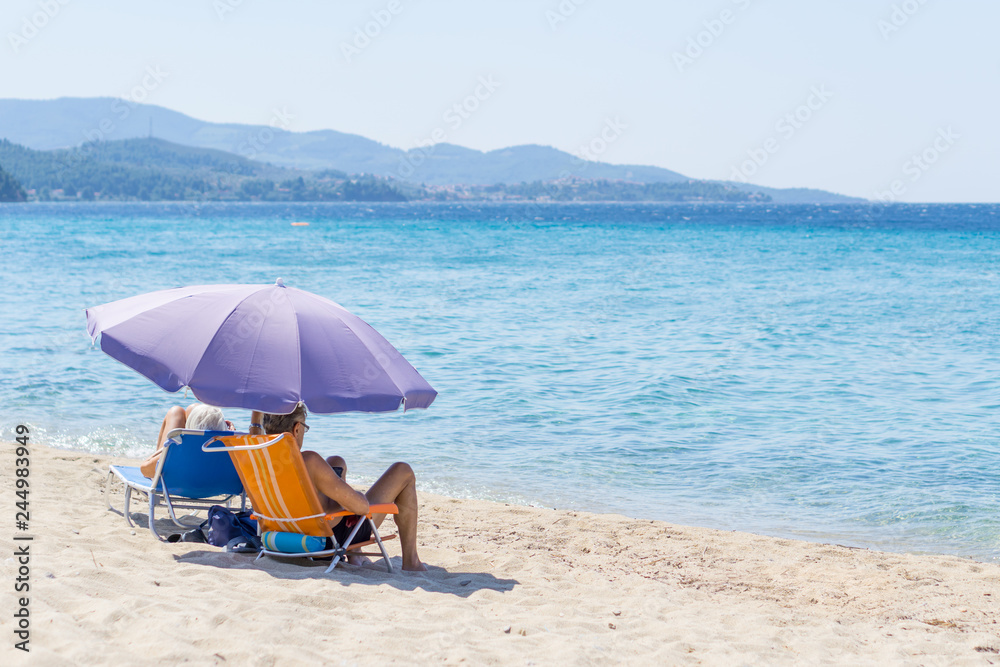 Man and woman sitting on small fold-out chairs on the beach under purple sunshade with a view of the blue sea water Man and woman sitting on small fold-out chairs on the beach under purple sunshade with a view of the blue sea water