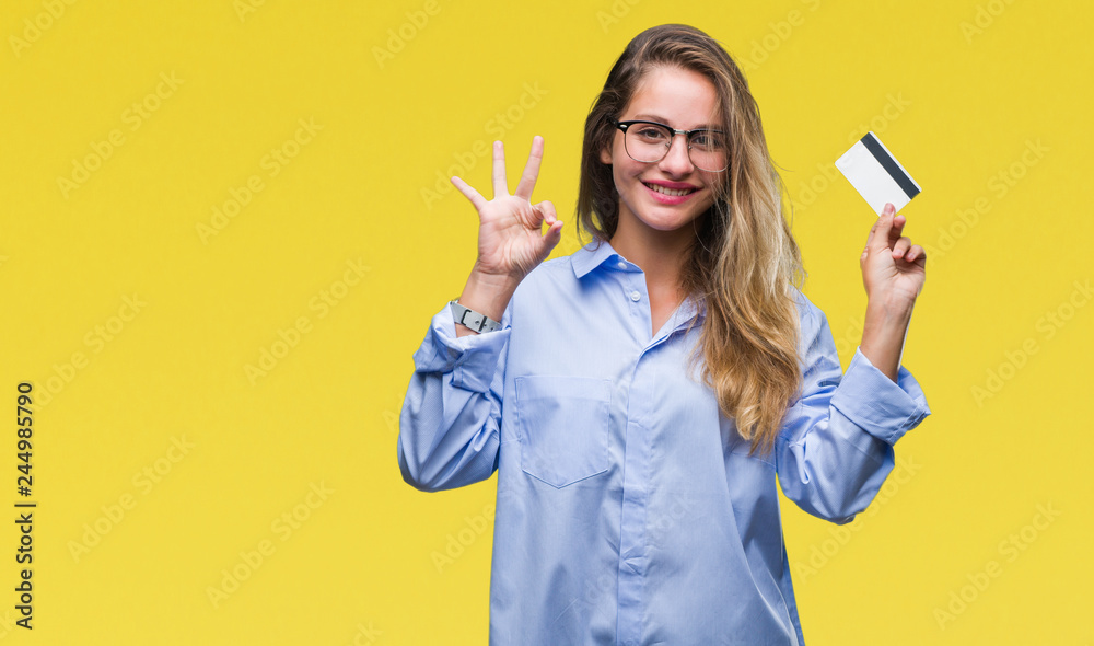 Young beautiful blonde woman holding credit card over isolated background doing ok sign with fingers, excellent symbol Young beautiful blonde woman holding credit card over isolated background doing ok sign with fingers, excellent symbol
