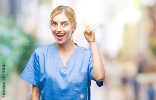 Young beautiful blonde doctor surgeon nurse woman over isolated background pointing finger up with successful idea. Exited and happy. Number one.