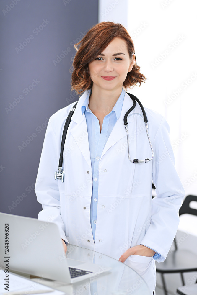Happy doctor woman at work. Portrait of female physician using laptop computer while standing near reception desk at clinic or emergency hospital. Medicine concept Happy doctor woman at work. Portrait of female physician using laptop computer while standing near reception desk at clinic or emergency hospital. Medicine concept