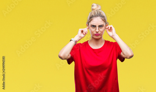 Young beautiful blonde woman wearing red t-shirt and glasses over isolated background covering ears with fingers with annoyed expression for the noise of loud music. Deaf concept.