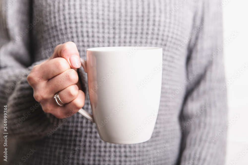 Female hand holding white cup in grey sweater background. Copy space Female hand holding white cup in grey sweater background. Copy space