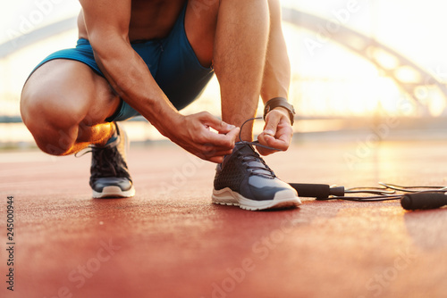 Fototapeta Naklejka Na Ścianę i Meble -  Close up of sporty man tying shoelace while kneeling in a court in the morning. Next to him skipping rope.