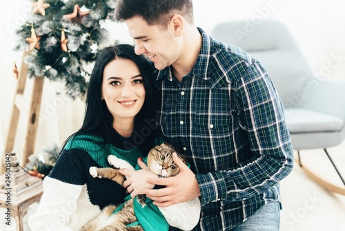 Couple in love sitting near Christmas tree and playing with cat at home