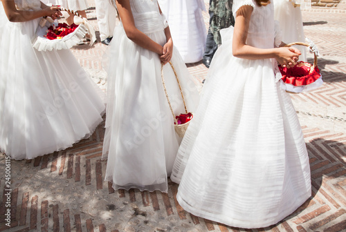 niñas llevando vestidos de comunión  blancos y largos con cesto con flores rojas photo