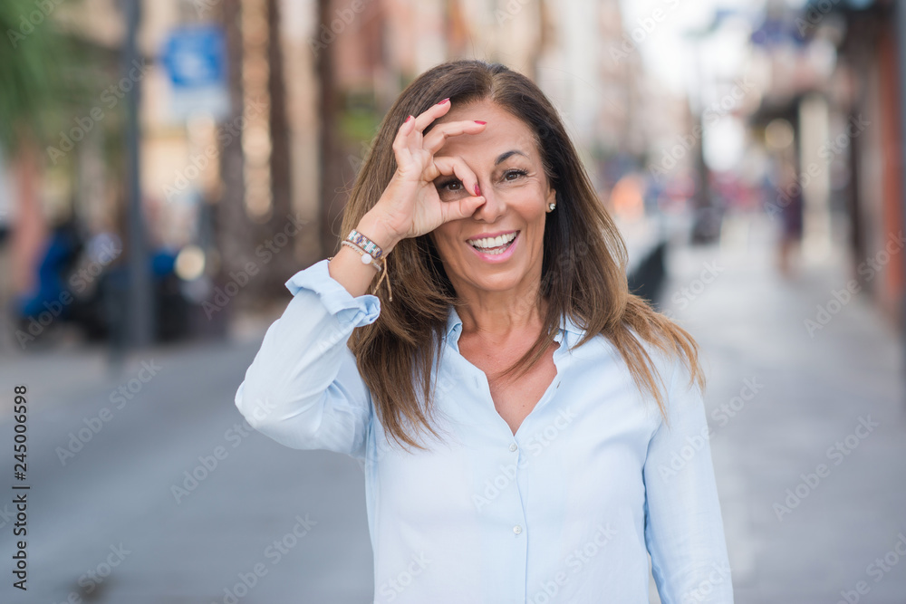 Beautiful middle age hispanic woman at the city street on a sunny day with happy face smiling doing ok sign with hand on eye looking through fingers