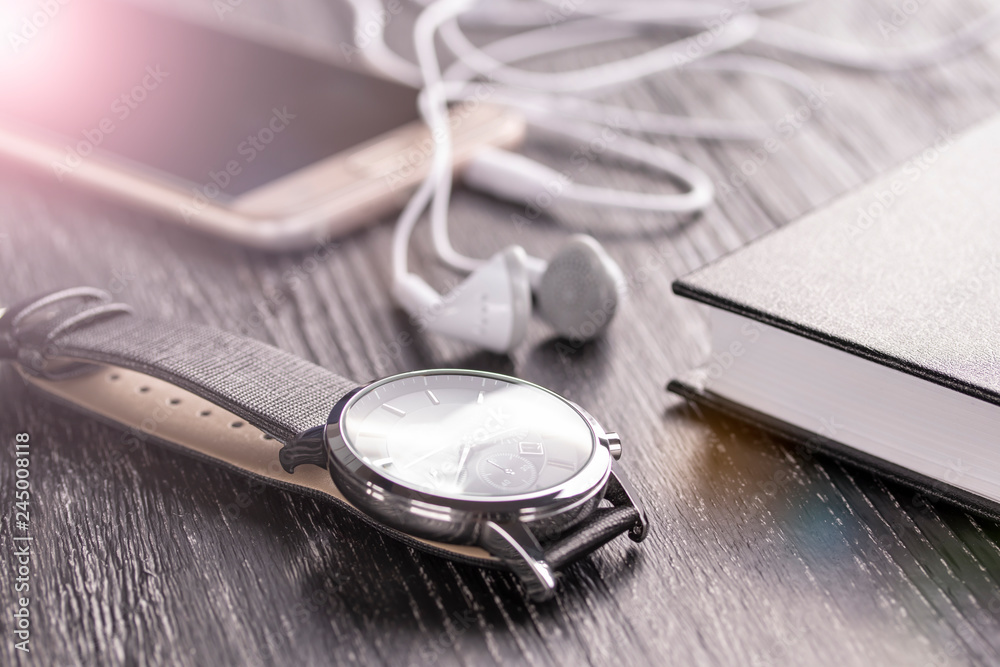 Wrist watch, mobile phone with headphones and notepad on an old dark office desktop Wrist watch, mobile phone with headphones and notepad on an old dark office desktop