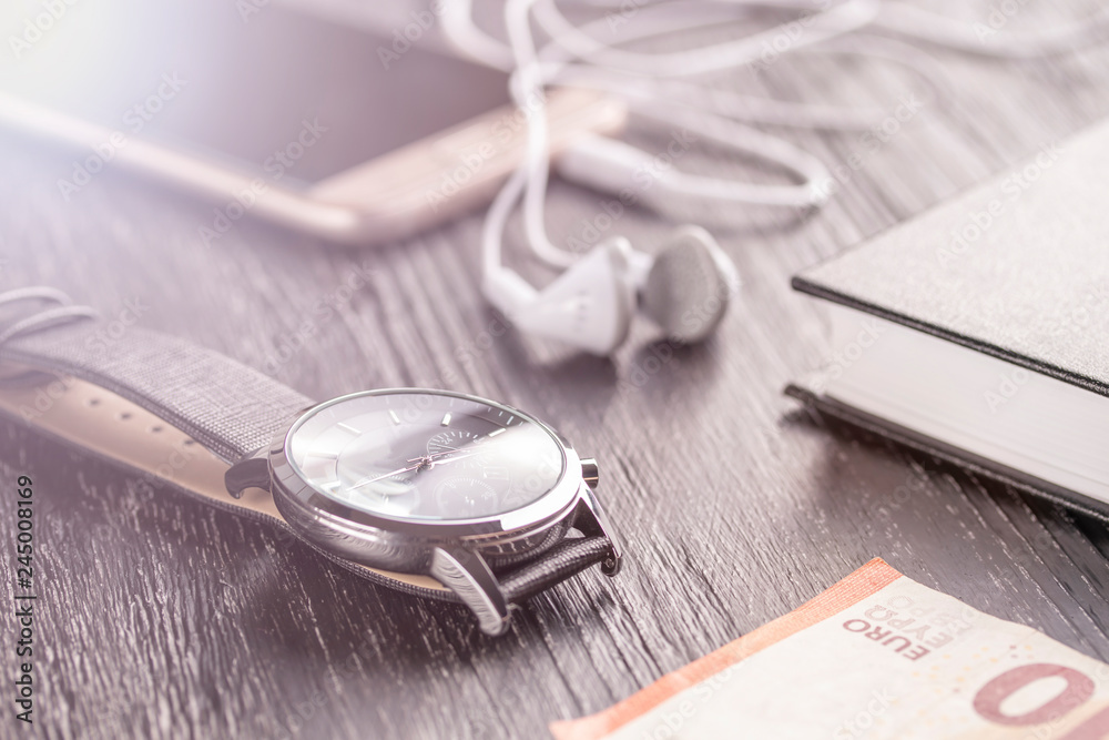 Wrist watch, mobile phone with headphones and notepad on the old dark office desktop. Nearby is the euro note Wrist watch, mobile phone with headphones and notepad on the old dark office desktop. Nearby is the euro note