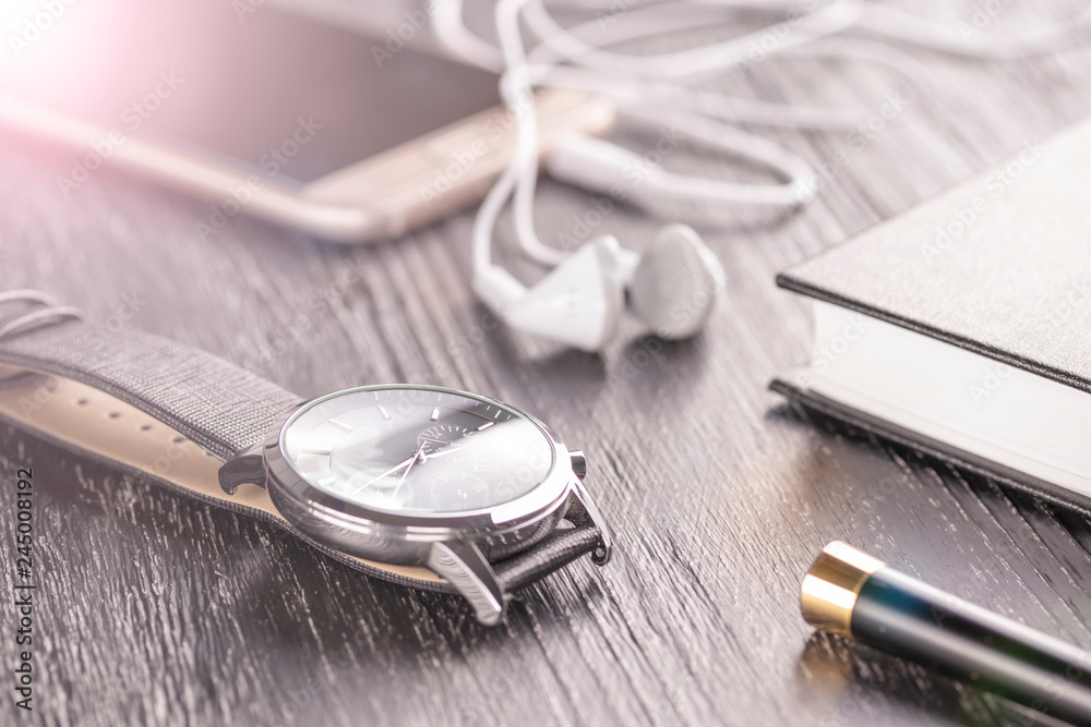 Wrist watch, mobile phone with headphones and a notepad with a pen on an old dark office desktop Wrist watch, mobile phone with headphones and a notepad with a pen on an old dark office desktop