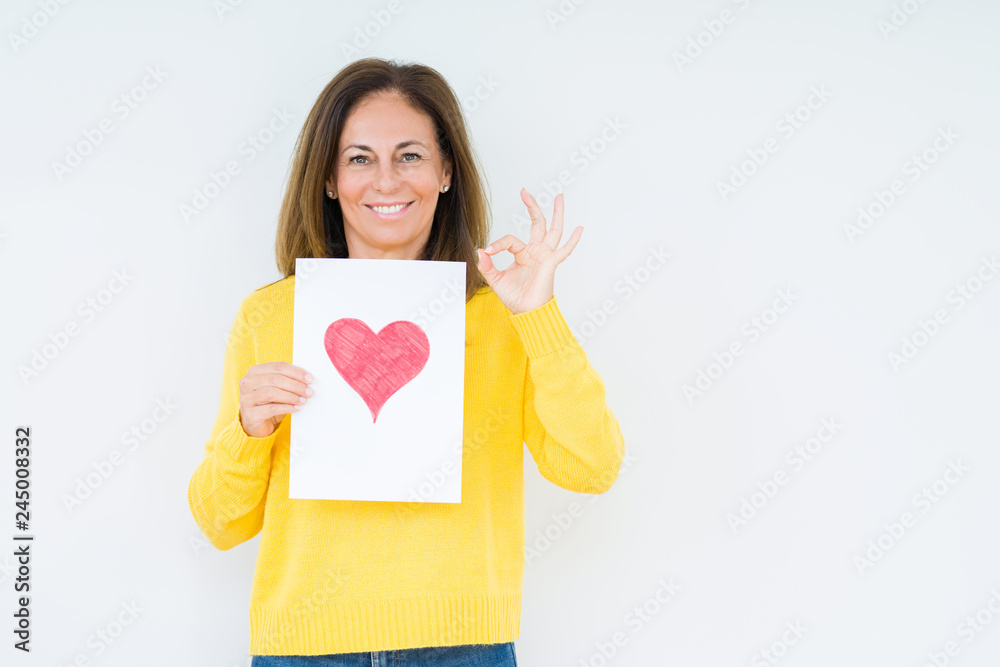 Middle age woman holding card gift with red heart over isolated background doing ok sign with fingers, excellent symbol Middle age woman holding card gift with red heart over isolated background doing ok sign with fingers, excellent symbol