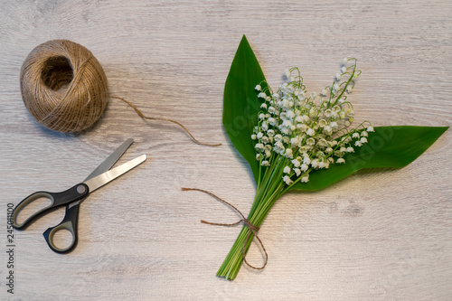 Bouquet of fresh lily of the valley flowers with skein of twine and scissors on light wooden background. Beautiful spring layout, top view